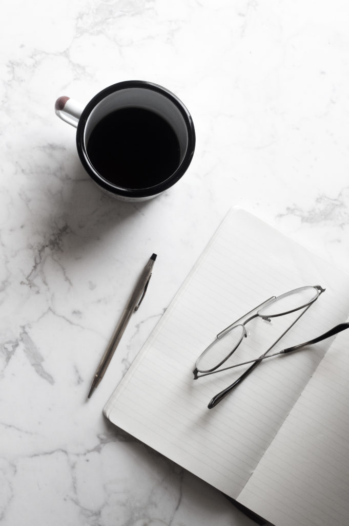 a photo from above of a cup of coffee, pen, paper and glasses on a white marble table